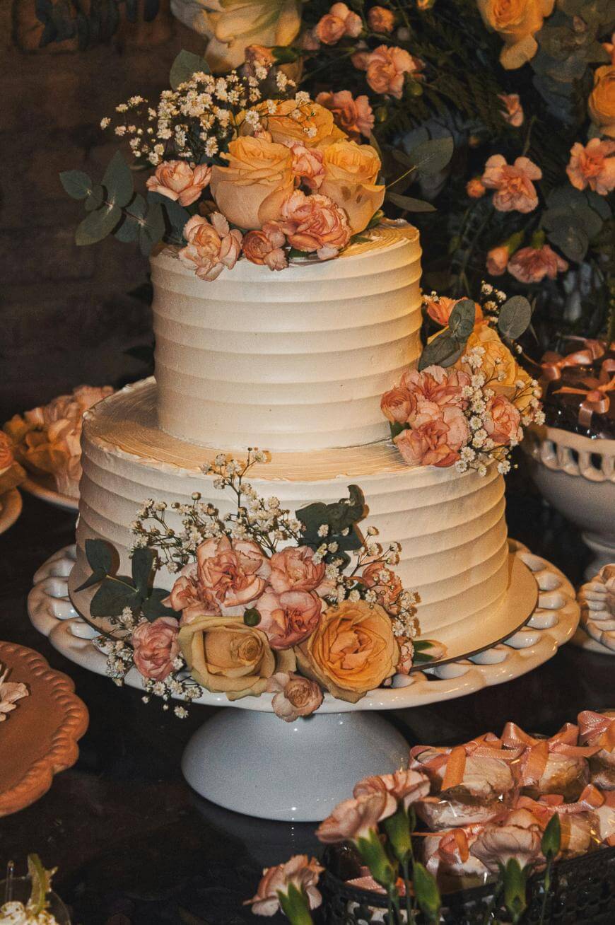 Two-tiered white cake decorated with peach and cream roses, small white flowers, and greenery, displayed on a white cake stand surrounded by matching floral arrangements.