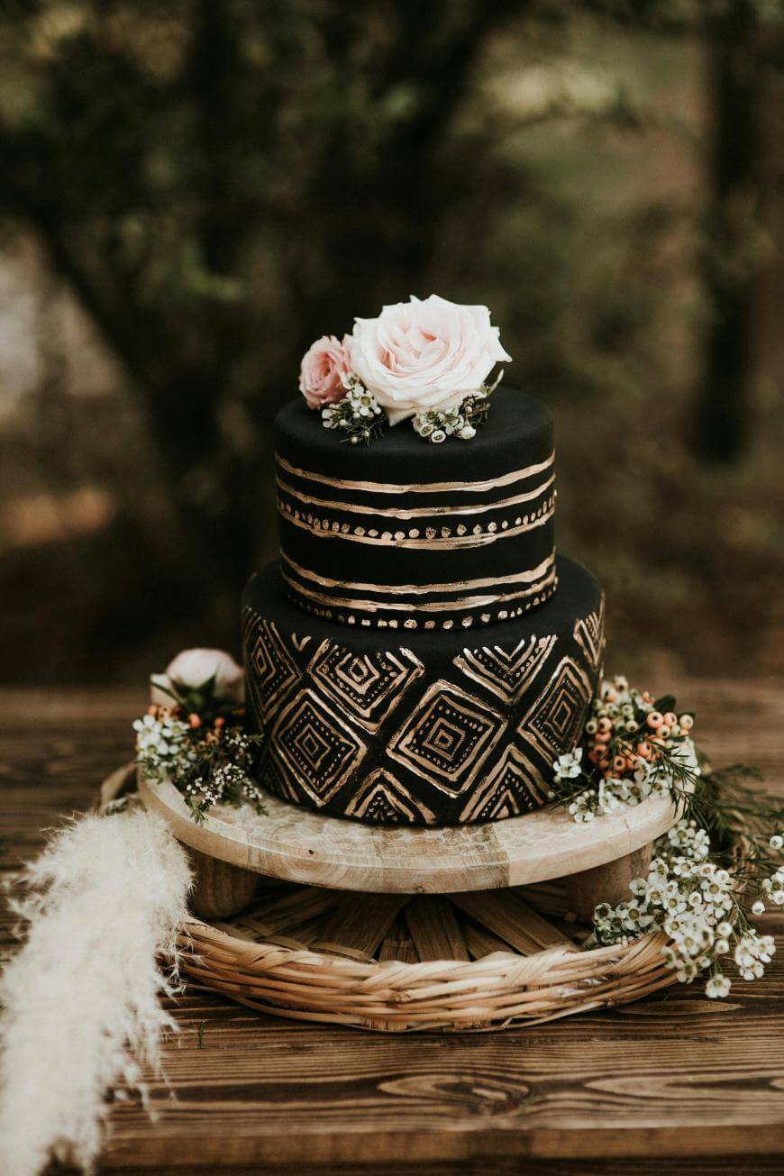 A two-tier black cake with gold geometric patterns and dots, topped with pink roses and greenery, sits on a rustic wooden stand, surrounded by flowers and foliage, outdoors in a natural setting.