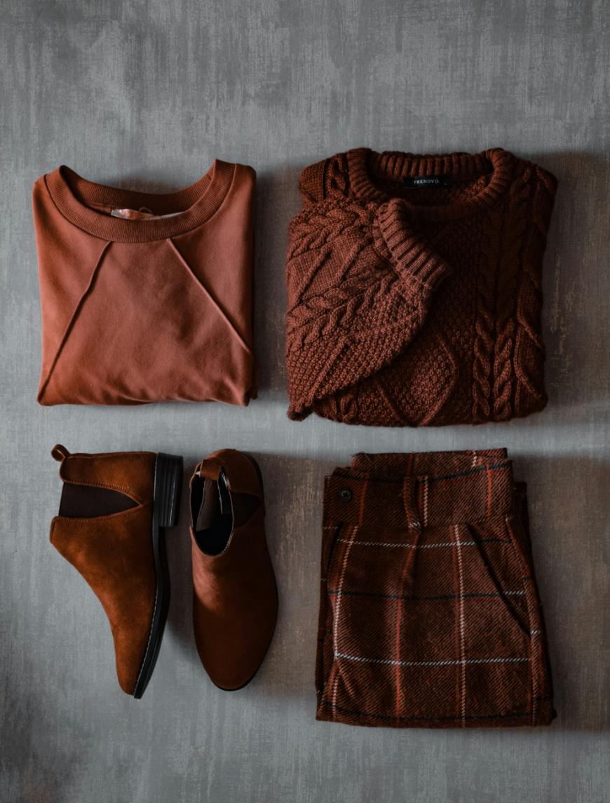 Four brown clothing items arranged neatly: a sweatshirt, a cable-knit sweater, a pair of ankle boots, and plaid shorts, all on a gray background.