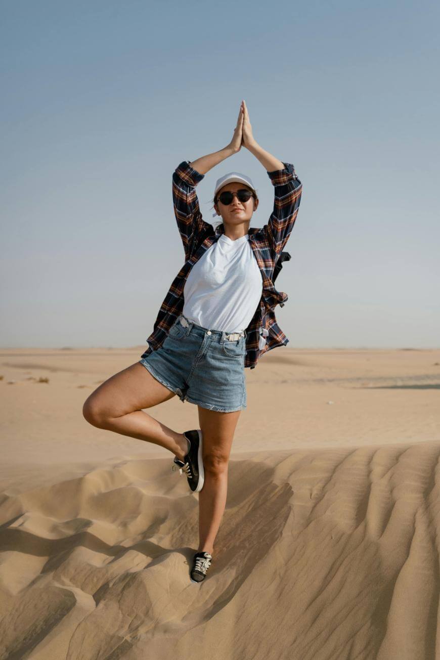 A person wearing sunglasses, a white t-shirt, plaid shirt, and denim shorts stands on one leg in a yoga pose on sandy dunes under a clear sky.