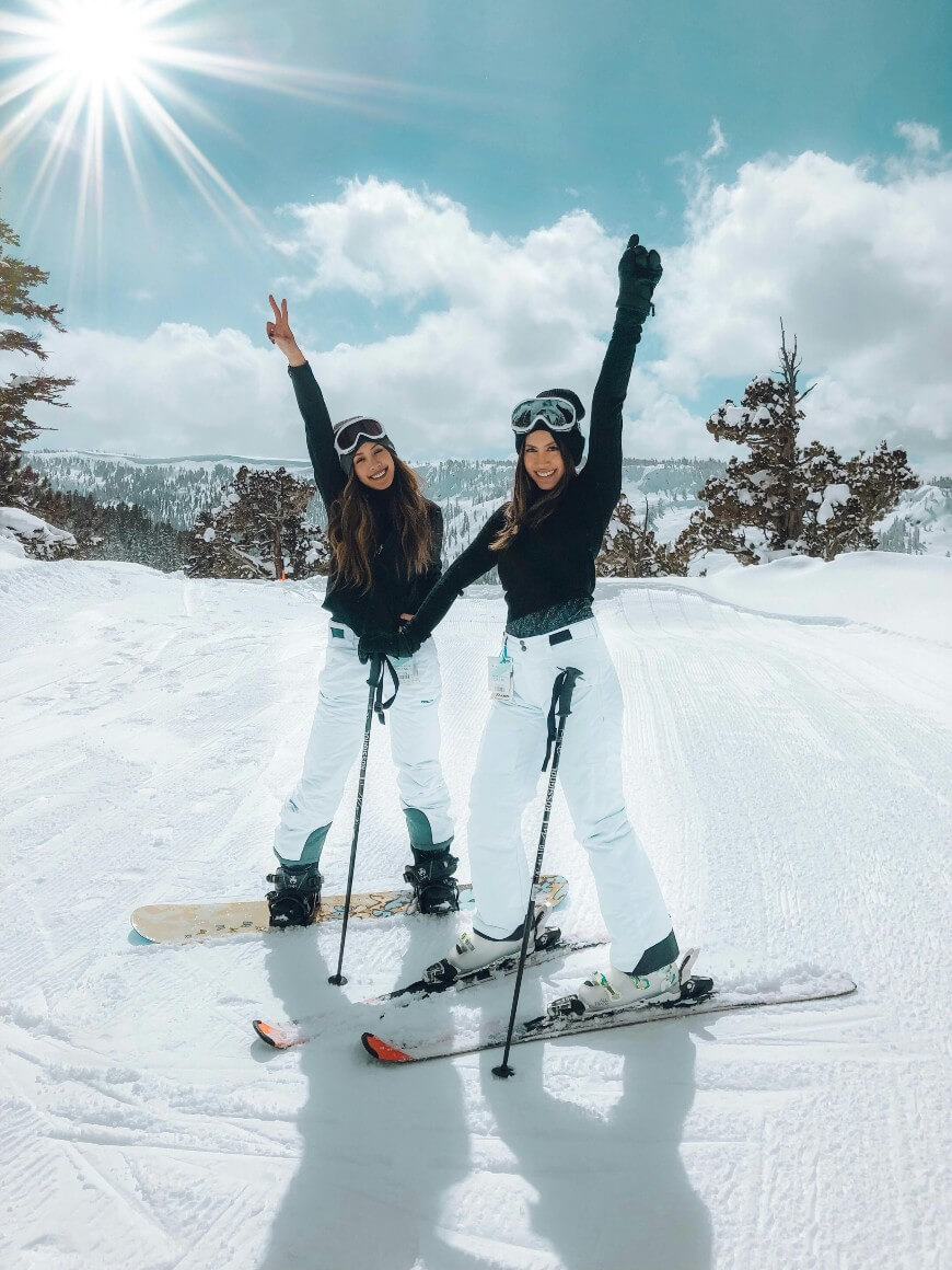 Two women in ski gear smile and pose on a snowy slope under a bright sun, with mountains and trees in the background; both are wearing goggles, black tops, and white ski pants, holding ski poles, enjoying a winter day.
