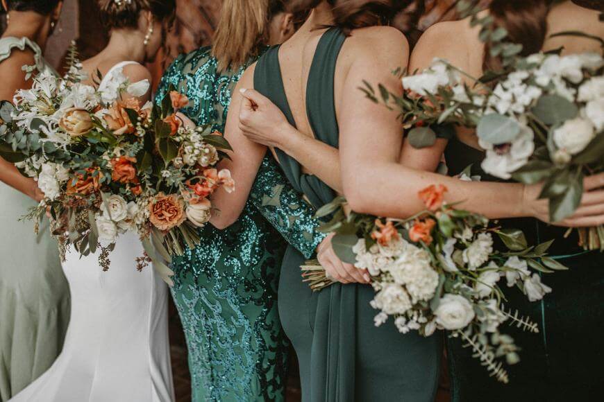 Group of women in green and white dresses stand close together, holding large bouquets of white, orange, and green flowers, with their arms linked around each other’s backs.