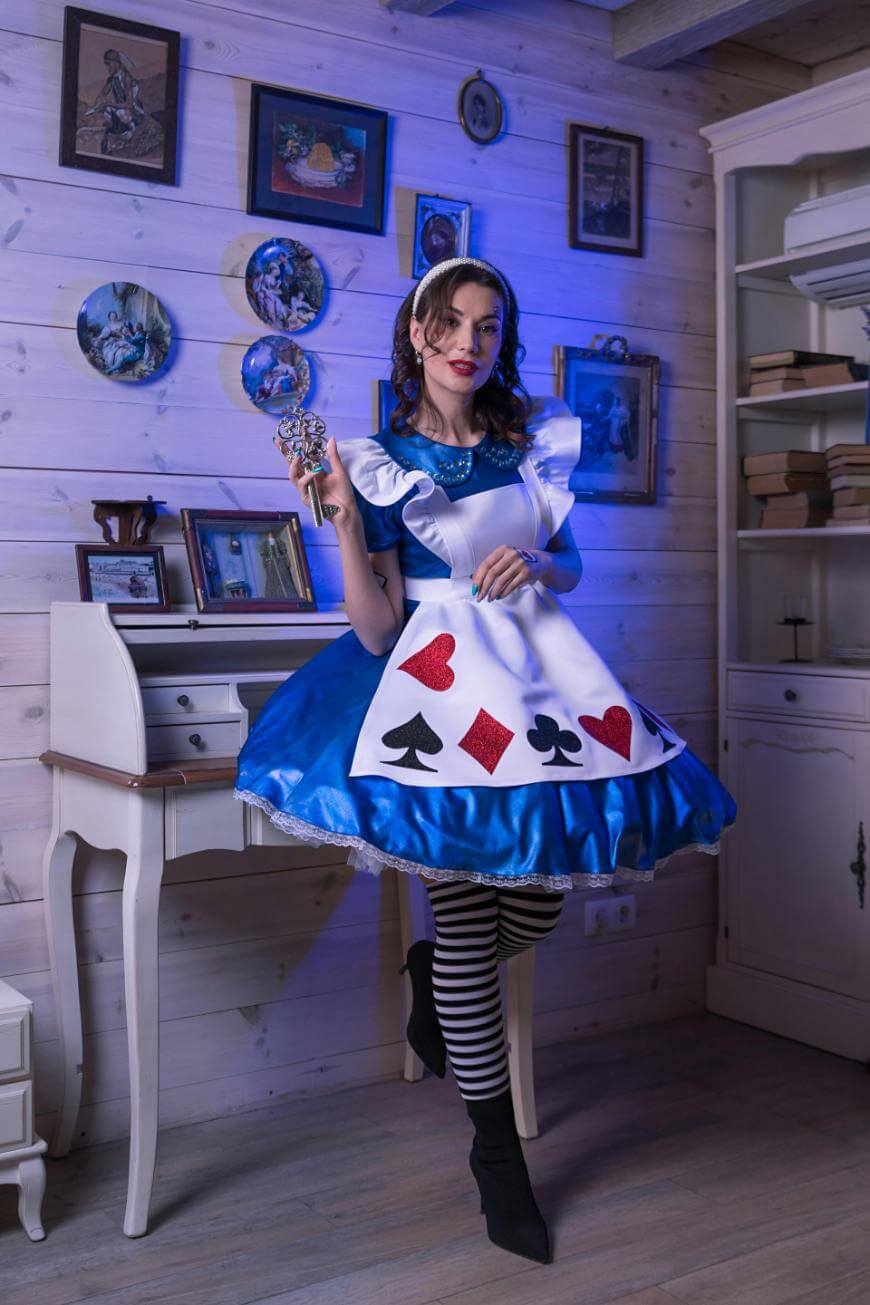 A woman dressed as Alice from Alice in Wonderland stands in a vintage room, wearing a blue dress with playing card symbols, striped tights, and holding a decorative key. Framed pictures and books decorate the cozy, softly-lit space.