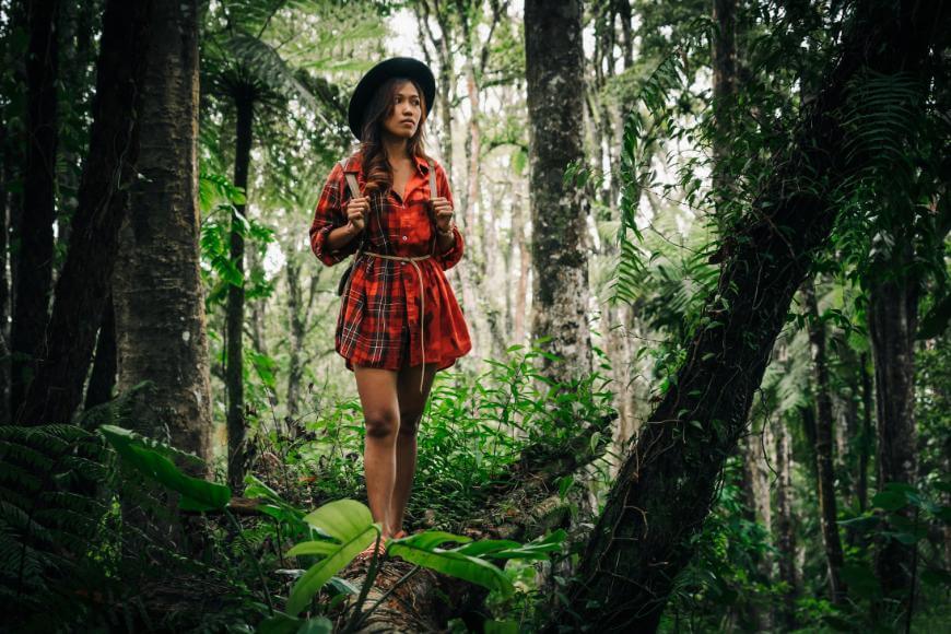A woman in a red plaid shirt, shorts, and a hat stands on a fallen tree with a backpack in a dense, green forest, surrounded by tall trees and lush foliage.