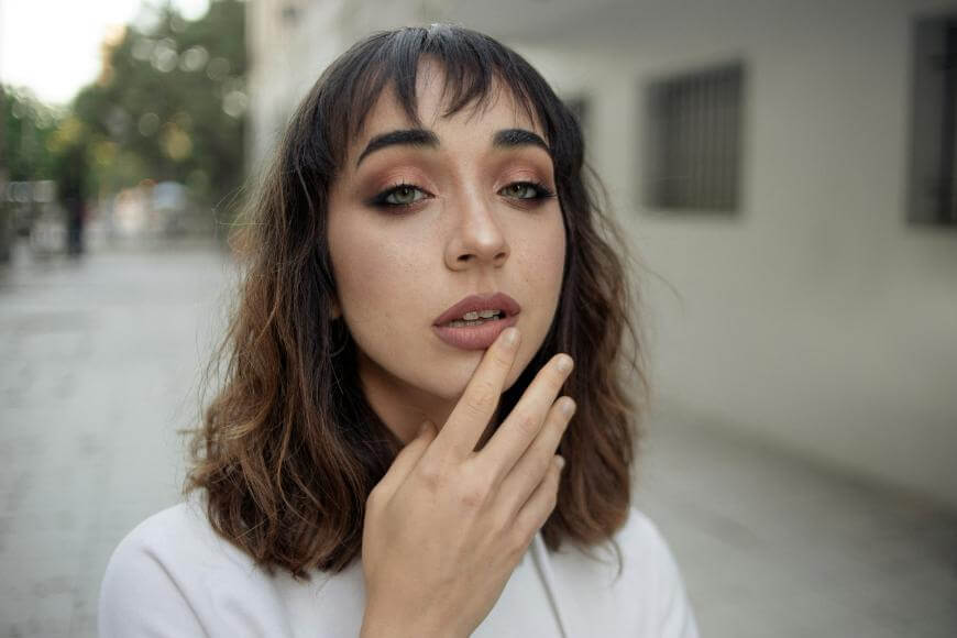 A person with shoulder-length brown hair and bangs gazes at the camera, touching their lips with one hand. They wear natural makeup and a white top, standing outdoors on a city street with blurred buildings in the background.