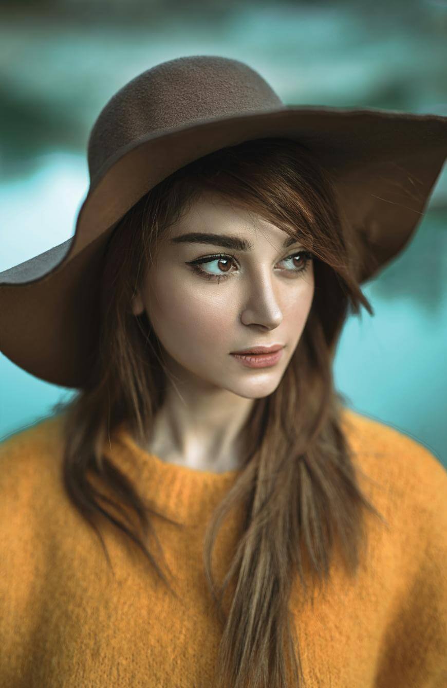 A young woman with long brown hair wearing a wide-brimmed brown hat and a mustard yellow sweater looks thoughtfully to the side, with a soft-focus blue and green background behind her.