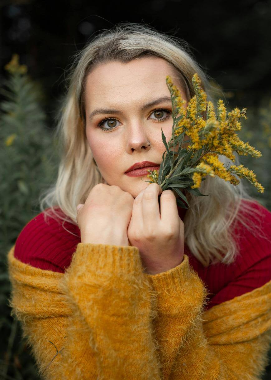 A woman with blonde hair and a nose ring holds yellow wildflowers near her face. She wears a red top and a fuzzy yellow sweater, gazing directly at the camera against a blurred outdoor background.