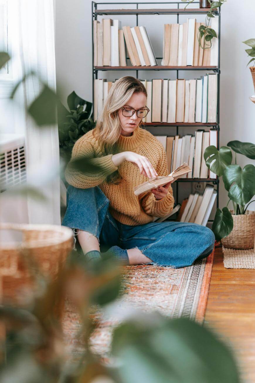 A woman with glasses sits on the floor in front of a bookshelf, reading a book. She wears a tan sweater and blue jeans, surrounded by houseplants in a cozy, sunlit room.