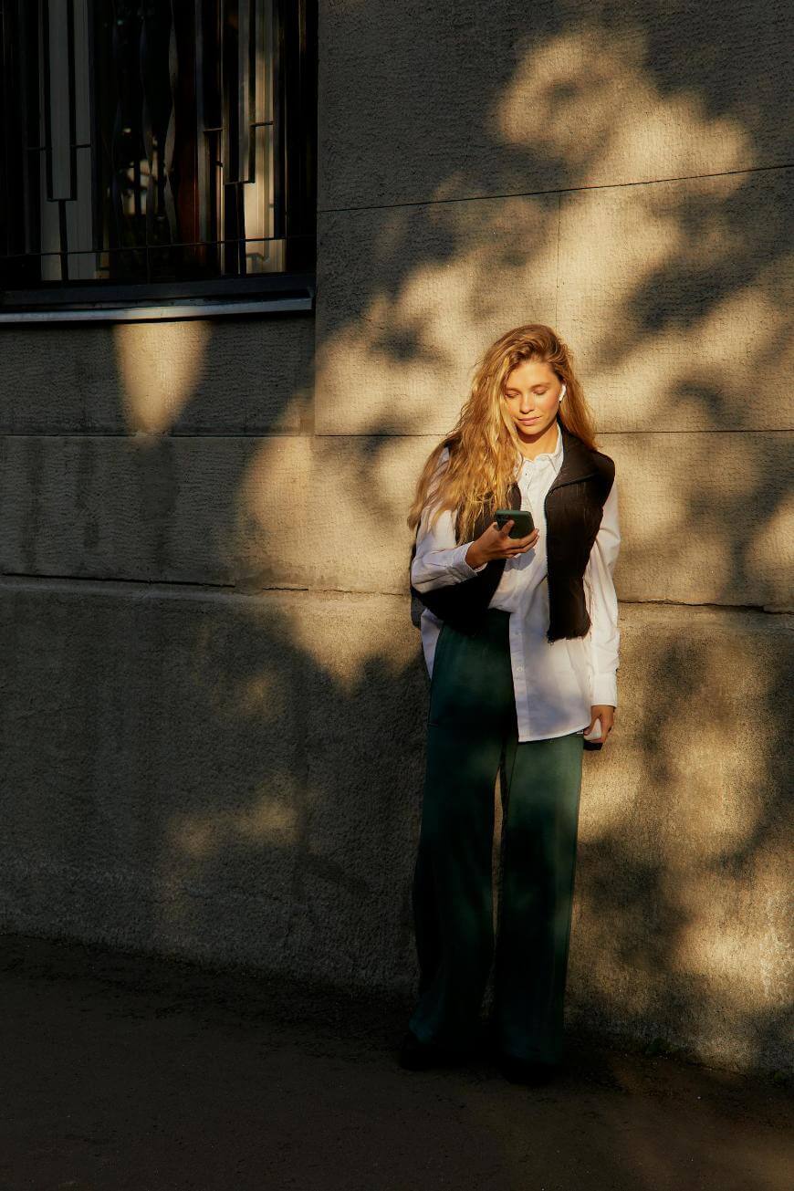 A woman with long blonde hair stands against a textured wall in warm sunlight, wearing a white shirt, dark vest, and dark pants, looking down at her phone with trees casting shadows on the wall.