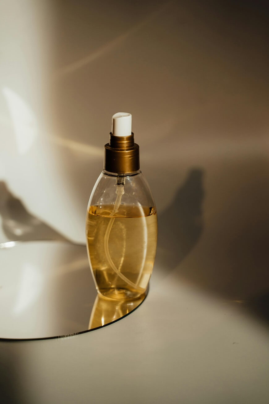 A clear spray bottle with a gold cap and light amber liquid inside is placed on a reflective surface, with soft light and shadows in the background.