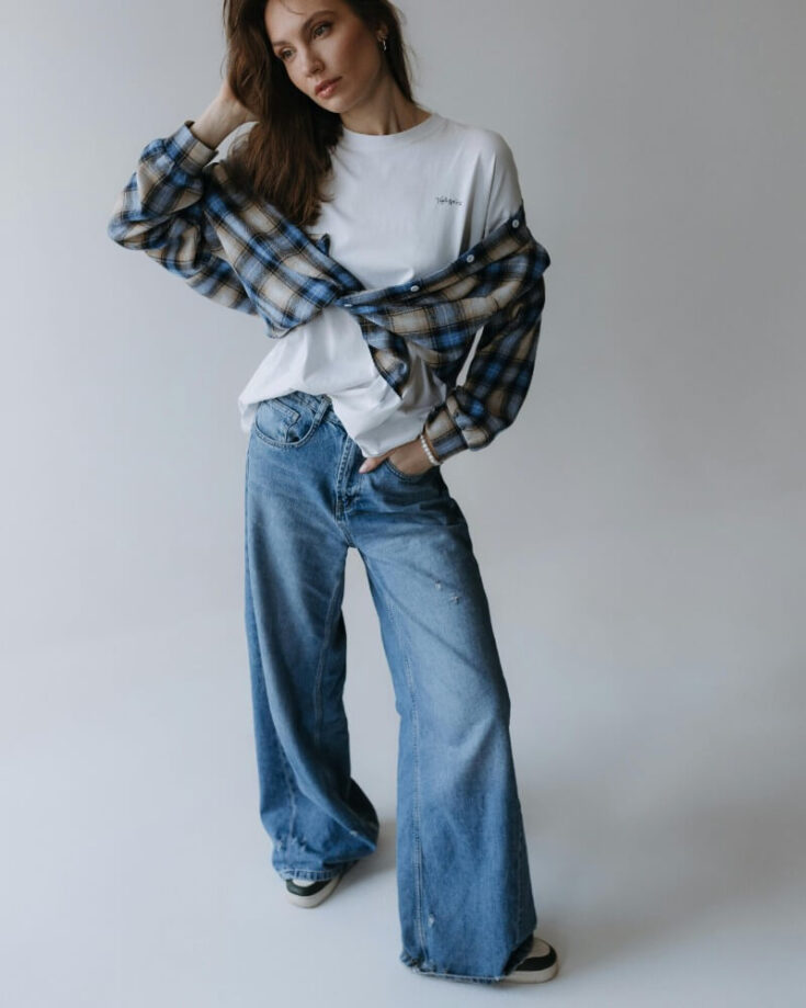A woman poses in wide-leg blue jeans, a white t-shirt, and a blue plaid shirt draped over her shoulders. She stands on a plain, light background with one hand touching her hair.
