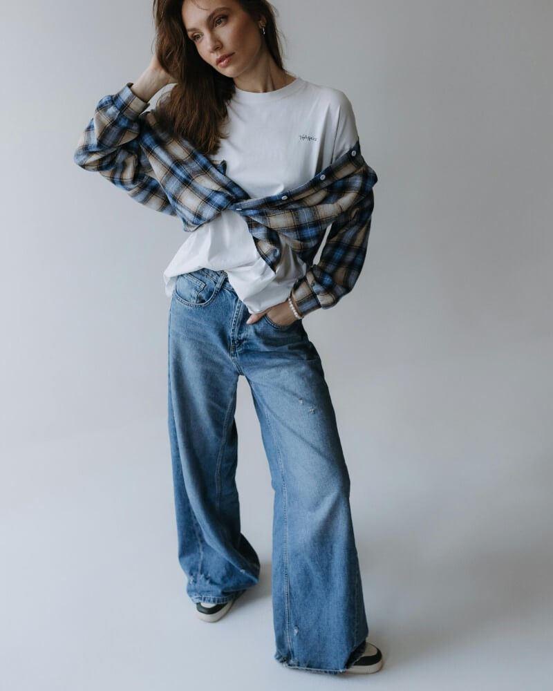A woman poses in wide-leg blue jeans, a white t-shirt, and a blue plaid shirt draped over her shoulders. She stands on a plain, light background with one hand touching her hair.