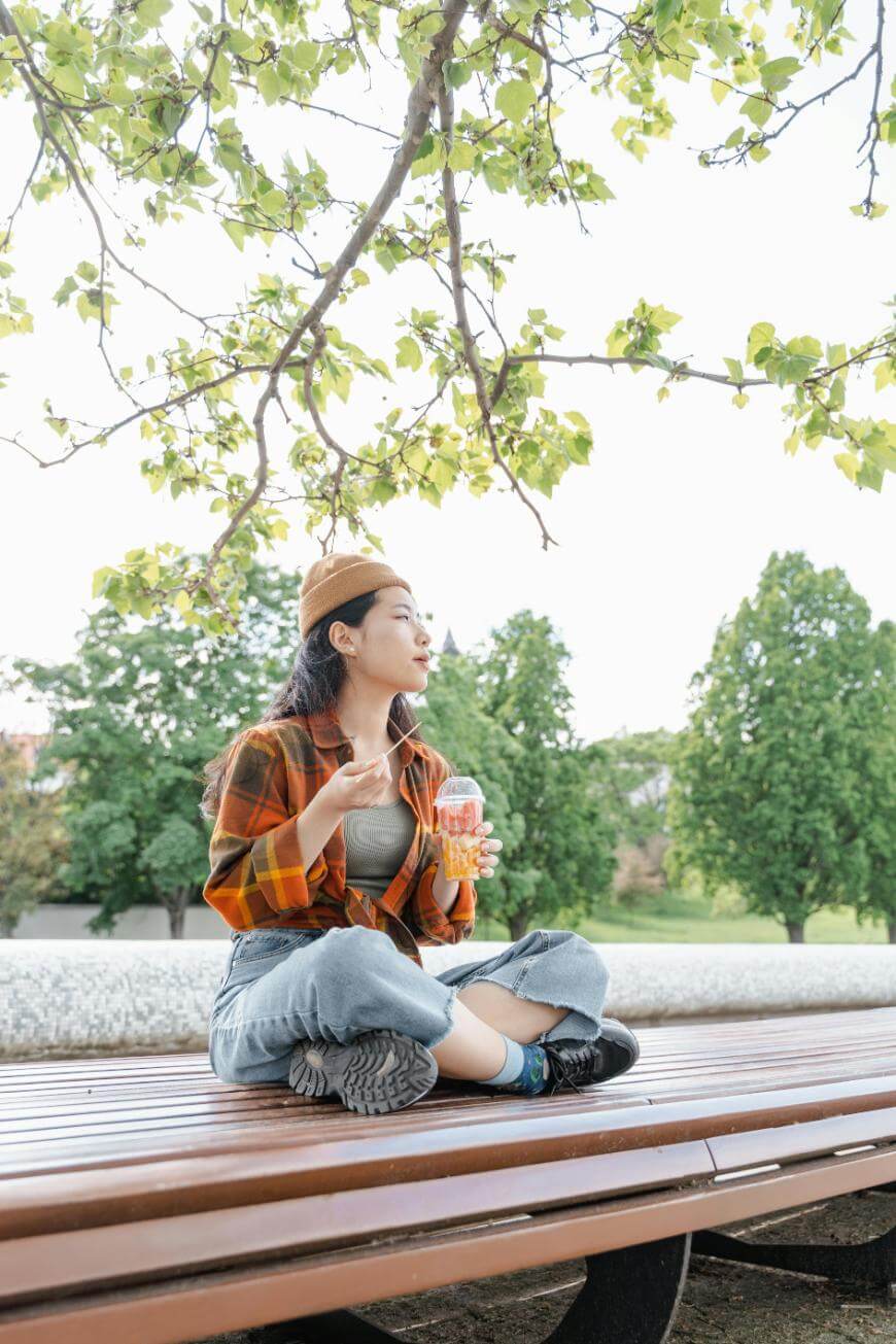 A young woman sits cross-legged on a wooden bench outdoors, eating from a cup with a spoon. She wears a plaid shirt, beanie, jeans, and sneakers. Green trees and leafy branches surround her in the background.