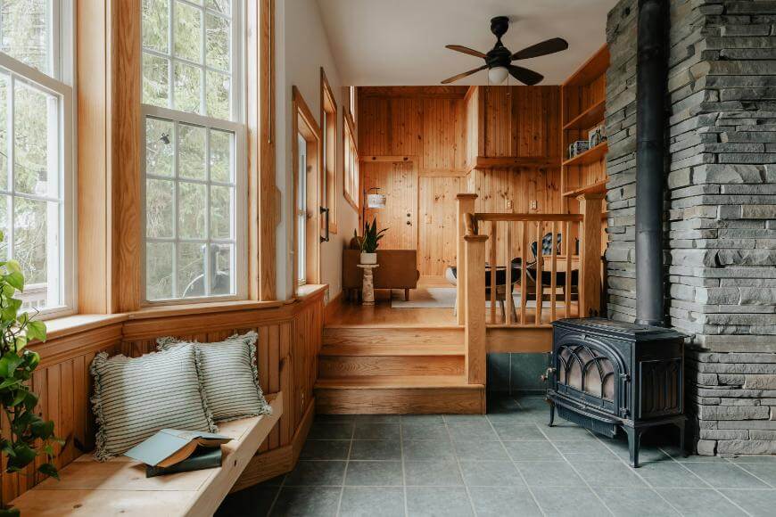 Cozy sunroom with large windows, a cushioned bench, a wood stove, stone and wood walls, green-tiled floor, and stairs leading to a living area with a ceiling fan and wooden shelves.