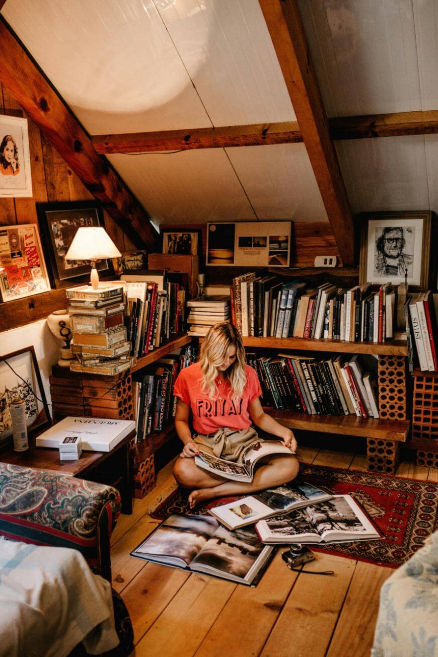 A woman with blonde hair sits cross-legged on a rug in a cozy attic room, reading a book. She is surrounded by bookshelves, framed photos, stacks of books, and warm wooden walls, creating an inviting reading nook.