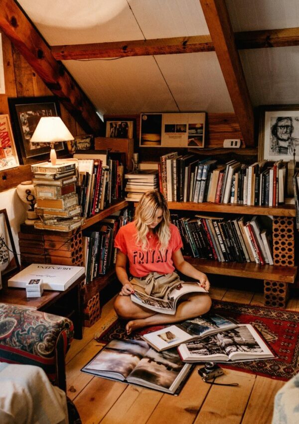 A woman sits cross-legged on a rug in a cozy attic library, reading a large book. She is surrounded by bookshelves, stacks of books, framed photos, and warm wooden beams. Natural light fills the inviting space.