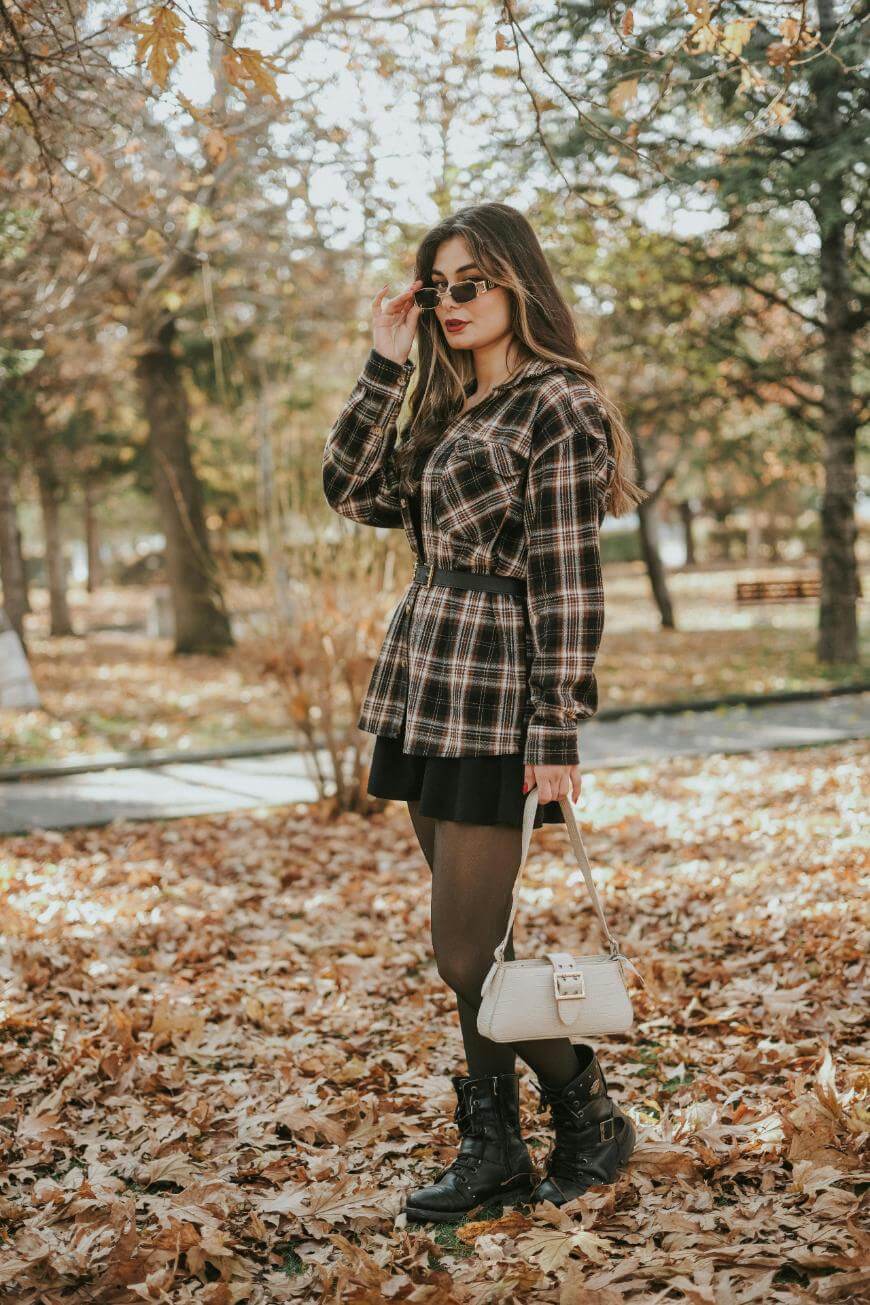 A woman stands on fallen autumn leaves in a park, wearing a plaid shirt jacket, black skirt, tights, black boots, sunglasses, and holding a beige handbag. Trees with brown leaves are visible in the background.