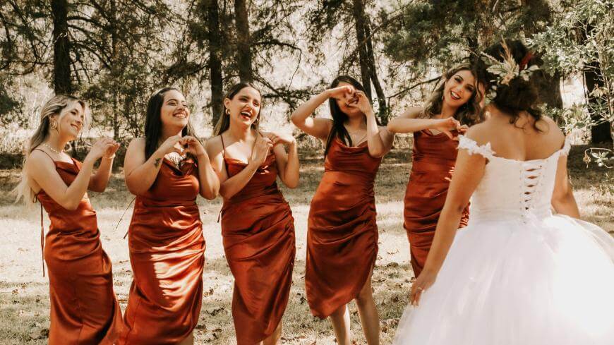 Five women in matching rust-colored dresses make heart shapes with their hands while smiling at a bride in a white gown, standing outdoors among trees on a sunny day.