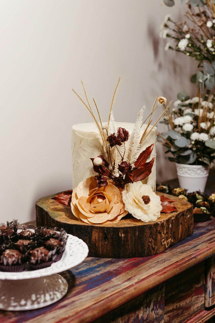 A rustic cake with white frosting, decorated with large dried flowers and foliage, sits on a wooden slab. Nearby, chocolate treats are displayed on a white stand. A vase with white flowers is in the background.