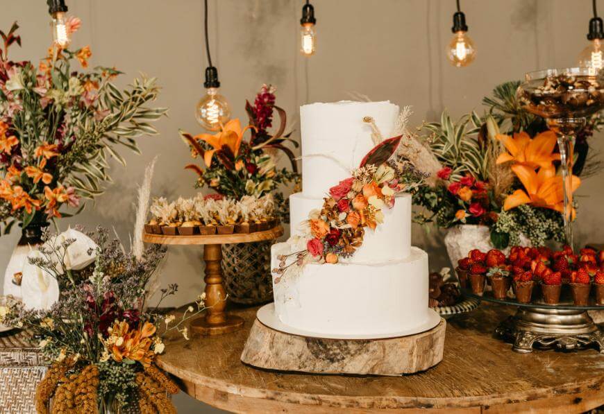 A white, three-tiered cake decorated with autumn flowers sits on a wooden stand. Surrounding it are floral arrangements, desserts, and strawberries. Hanging Edison bulbs light the rustic display.