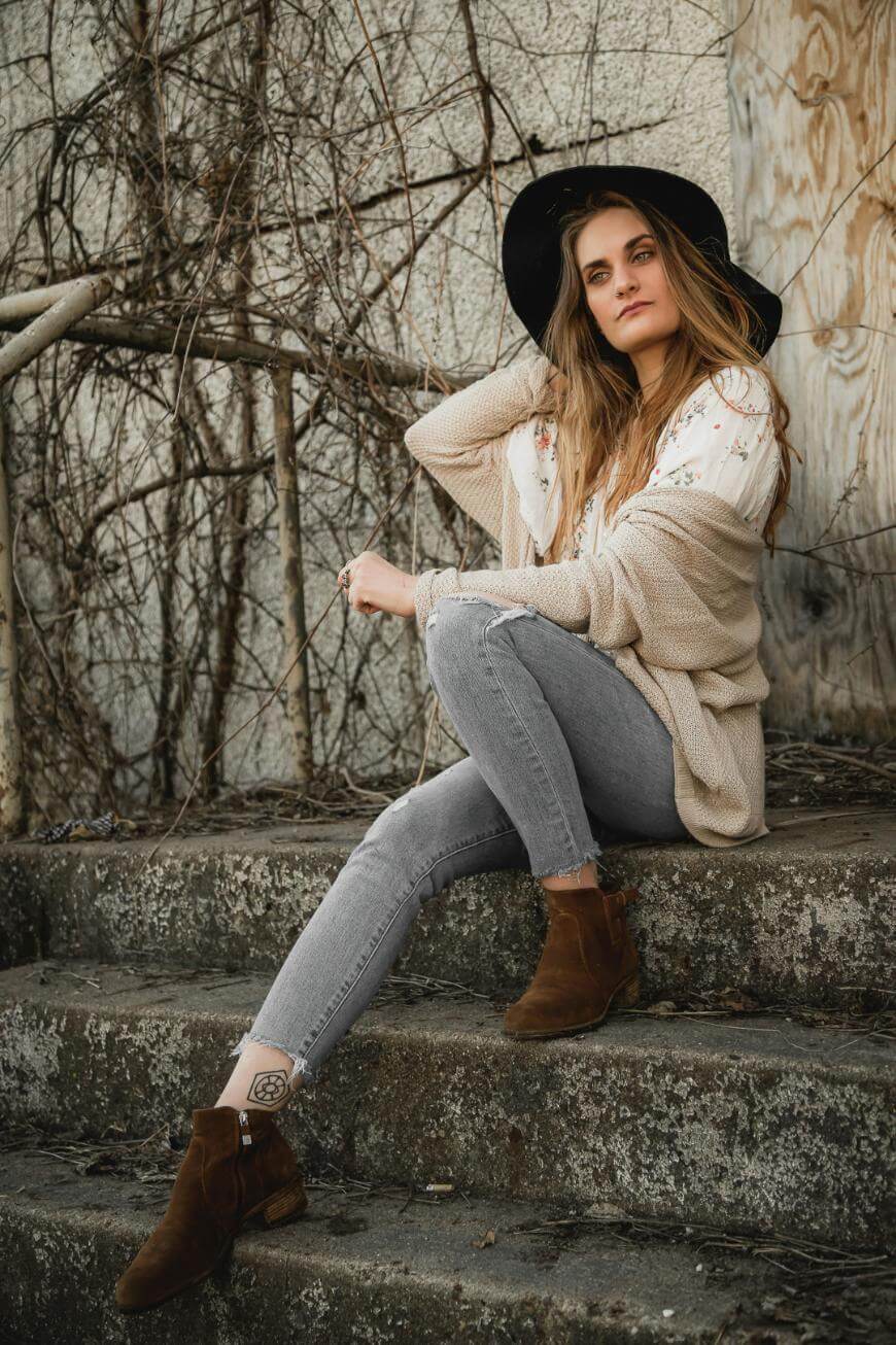 A woman wearing a black hat, light sweater, floral blouse, gray jeans, and brown boots sits on outdoor concrete steps against a textured wall with bare vines, looking thoughtfully at the camera.