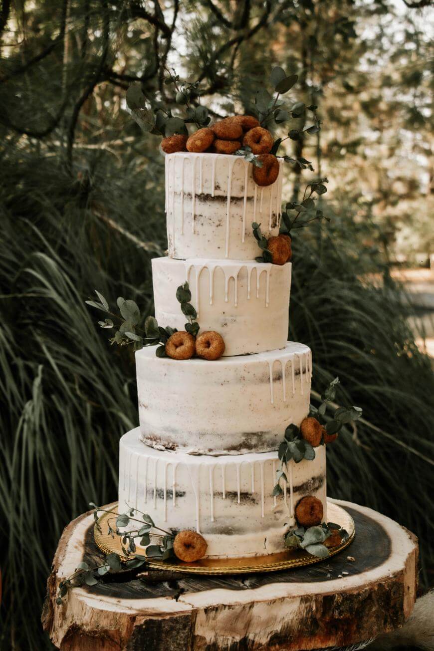 A four-tiered rustic wedding cake with white drip icing, decorated with small donuts and greenery, displayed on a wooden slice outdoors with trees and foliage in the background.
