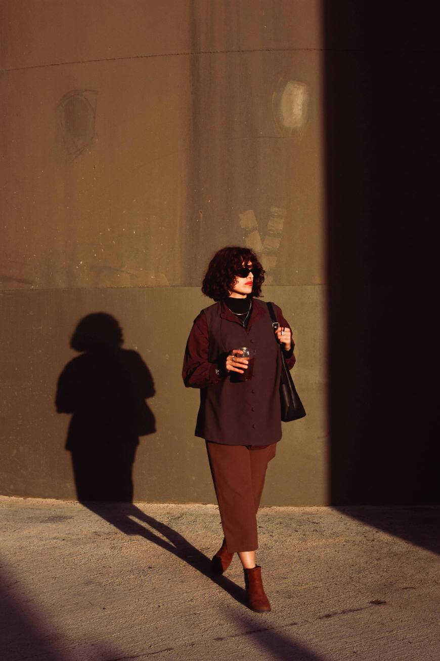 A woman with curly hair and sunglasses walks in sunlight, casting a shadow on a brown wall. She wears a dark outfit, holds a drink in one hand, and carries a black bag in the other.