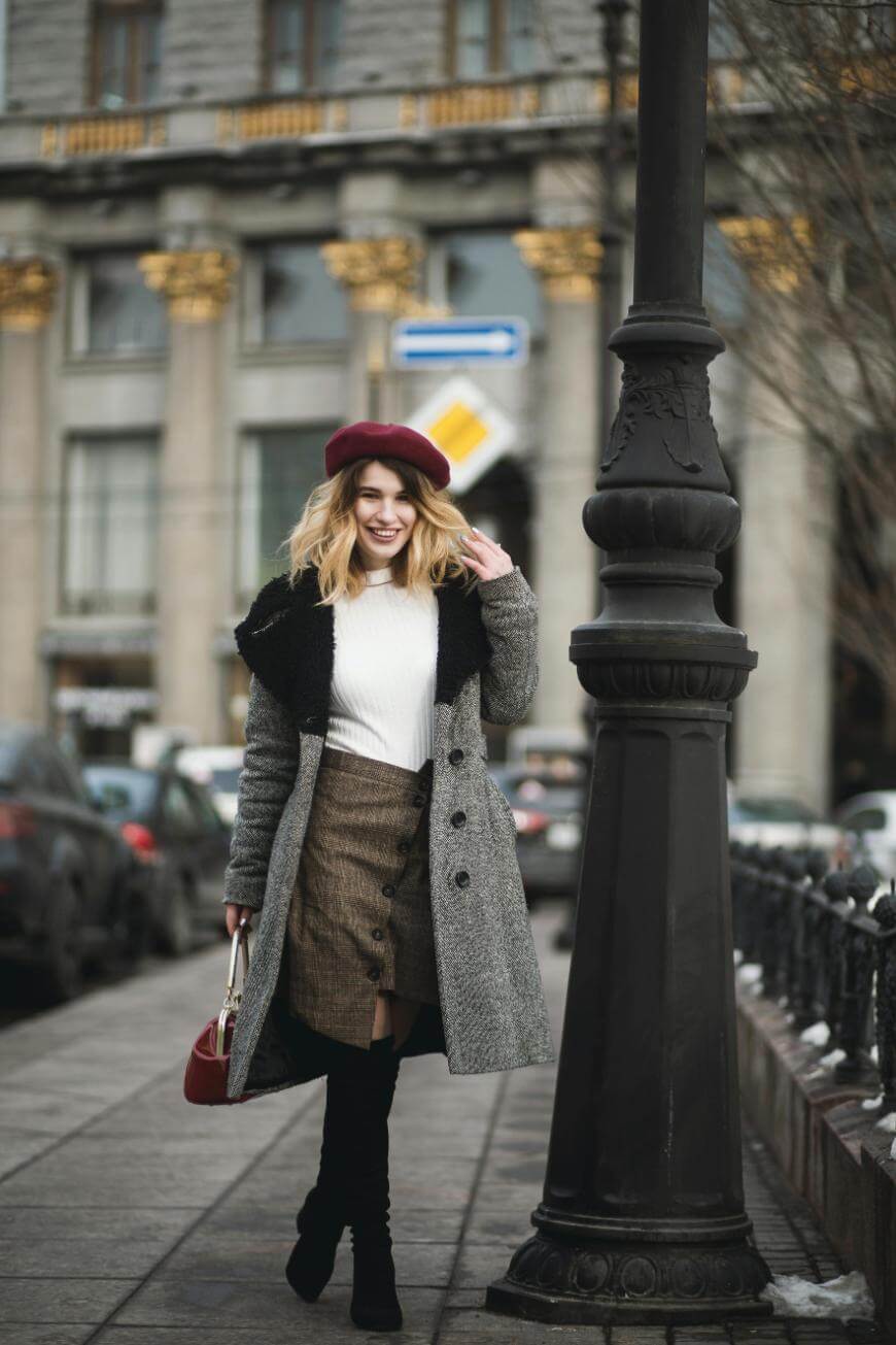 A woman wearing a gray coat, white sweater, brown skirt, and red beret smiles while walking on a city sidewalk. She holds a small red handbag and there are parked cars and a building in the background.
