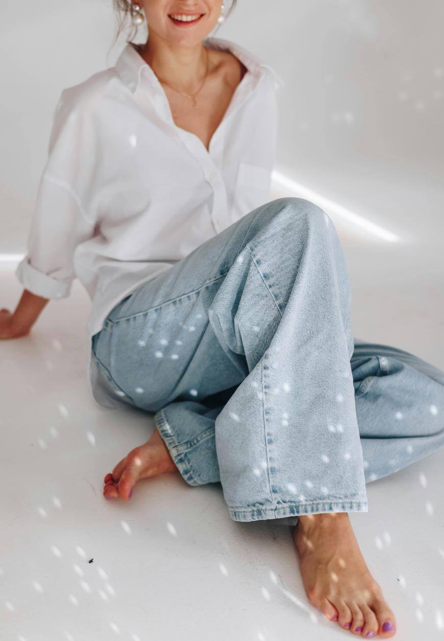 A woman in a white button-up shirt and light blue jeans sits barefoot on the floor, smiling softly, with scattered light spots reflecting on her clothes and the white background.