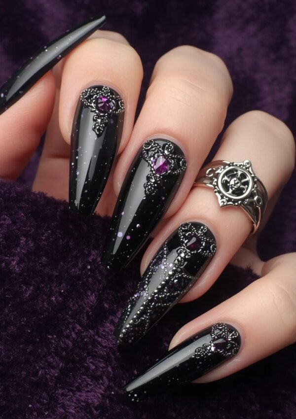 A hand with long, glossy black Witchy Nails decorated with silver filigree and purple gemstones rests on a purple velvet fabric. The person wears a silver ring with a decorative design.