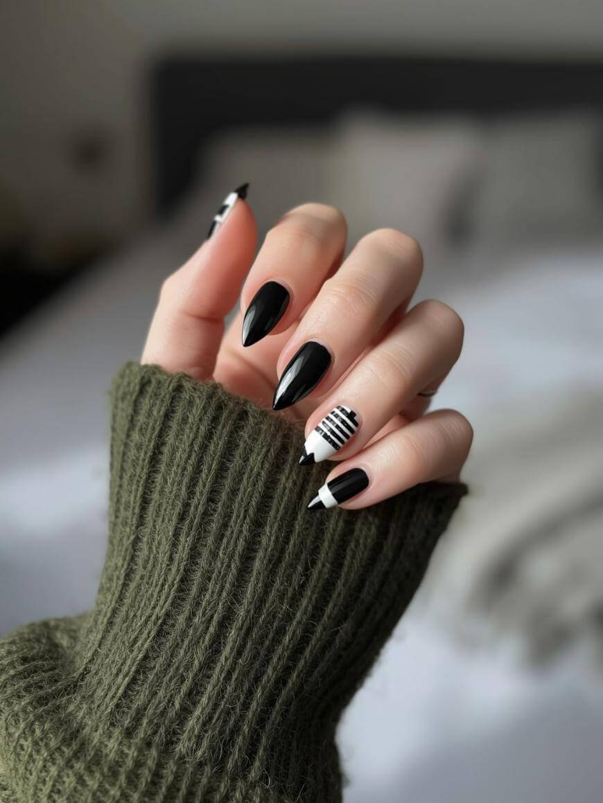 A hand with almond-shaped nails painted in black and white geometric designs, including stripes and triangles, is shown. The person is wearing a green ribbed sweater, and a blurred bed is in the background.