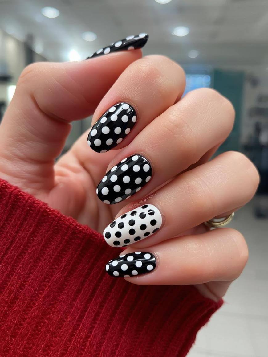 A hand with manicured nails featuring a black and white polka dot design; most nails are black with white dots, while the ring finger is white with black dots. The person is wearing a red sweater.