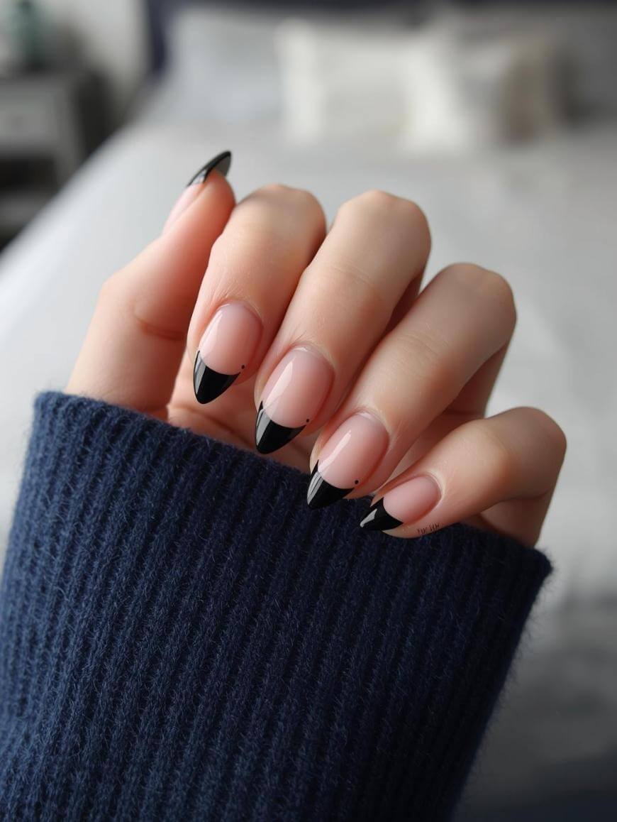 A hand with long, almond-shaped nails featuring a black French tip design and small black dots, shown against a blurred background. The hand is partially covered by a dark blue ribbed sweater sleeve.