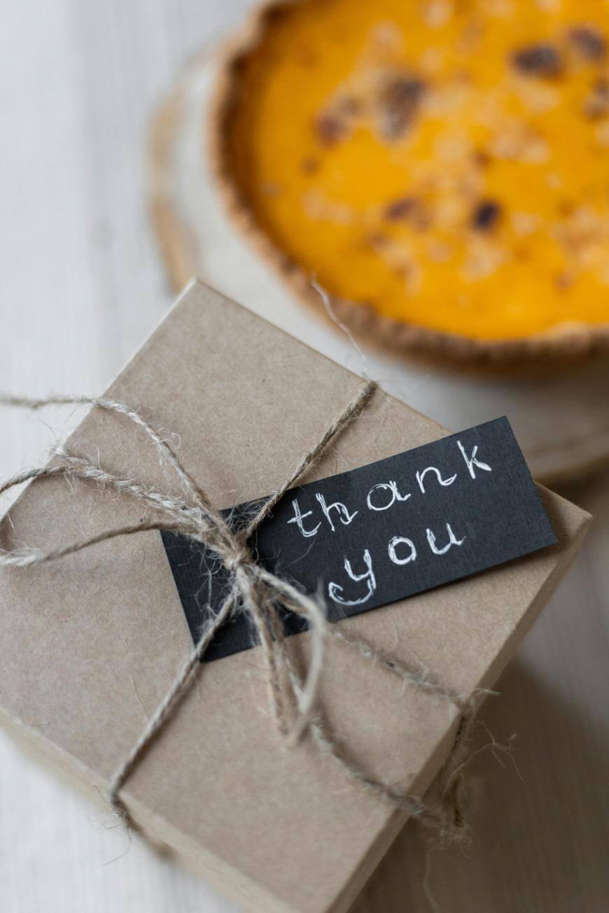 A small brown gift box tied with twine and a black tag that says “thank you” in white handwriting, placed on a white surface next to a blurred pie in the background.