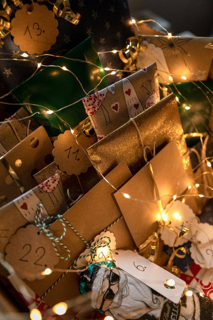 A close-up of various numbered brown paper packages, some with festive string and decorative tags, surrounded by warm white fairy lights, creating a cozy holiday atmosphere.