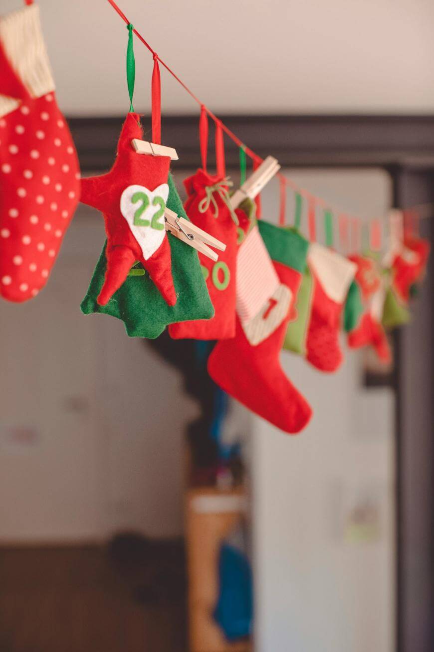 A string of colorful felt Christmas stockings and pouches, some with numbers, hangs on a red ribbon with clothespins, used as an advent calendar. The background is blurred indoors.