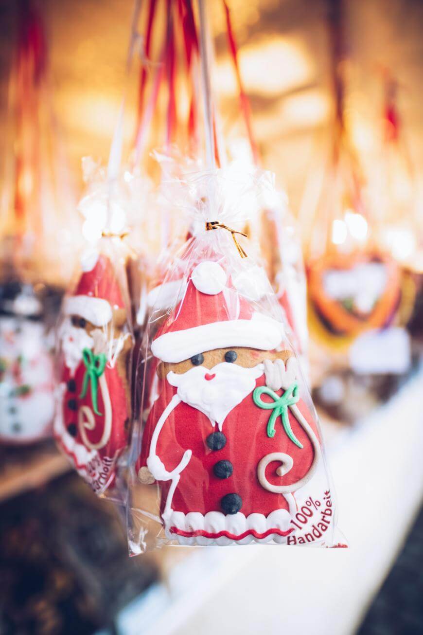 A decorated gingerbread cookie shaped like Santa Claus, wrapped in clear plastic and tied with a gold twist, hangs on display with other festive treats in the background.