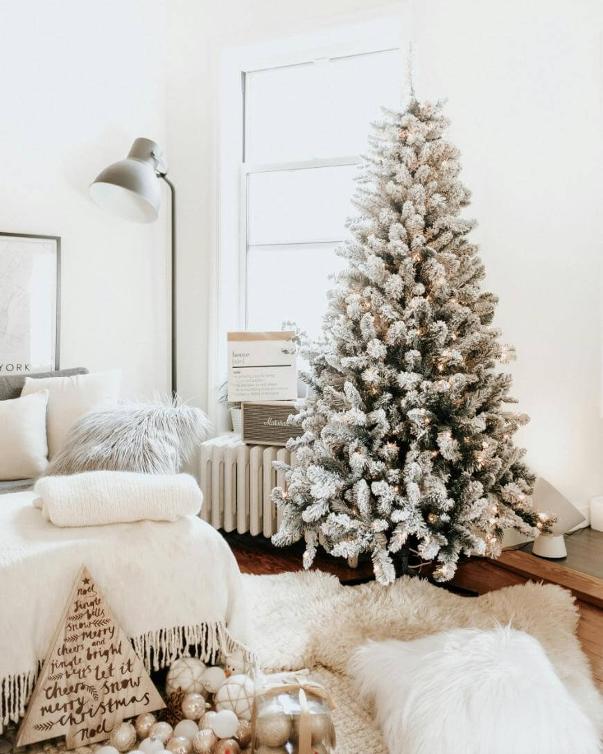 A cozy living room with a flocked Christmas tree decorated with white lights, surrounded by neutral-toned pillows, a fluffy rug, a white throw, a basket of ornaments, and soft ambient lighting.