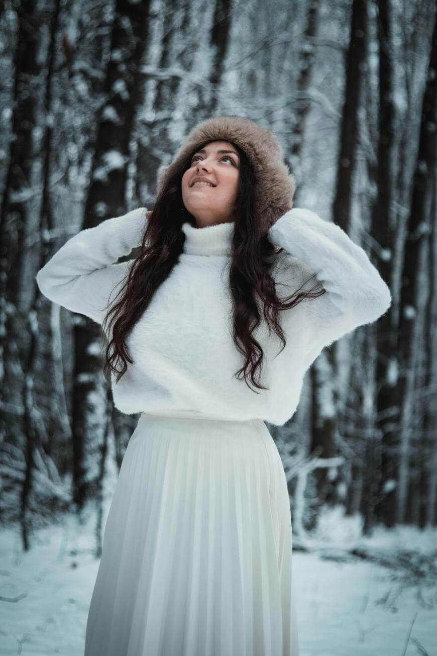 A woman wearing a white turtleneck sweater, long white skirt, and a furry hat stands in a snowy forest, looking up and smiling with her hands on her hat. Snow covers the ground and trees in the background.
