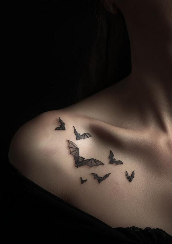 A close-up of a persons shoulder and collarbone with a tattoo of several black bats in flight. The background is dark, and the person is wearing an off-the-shoulder black top.