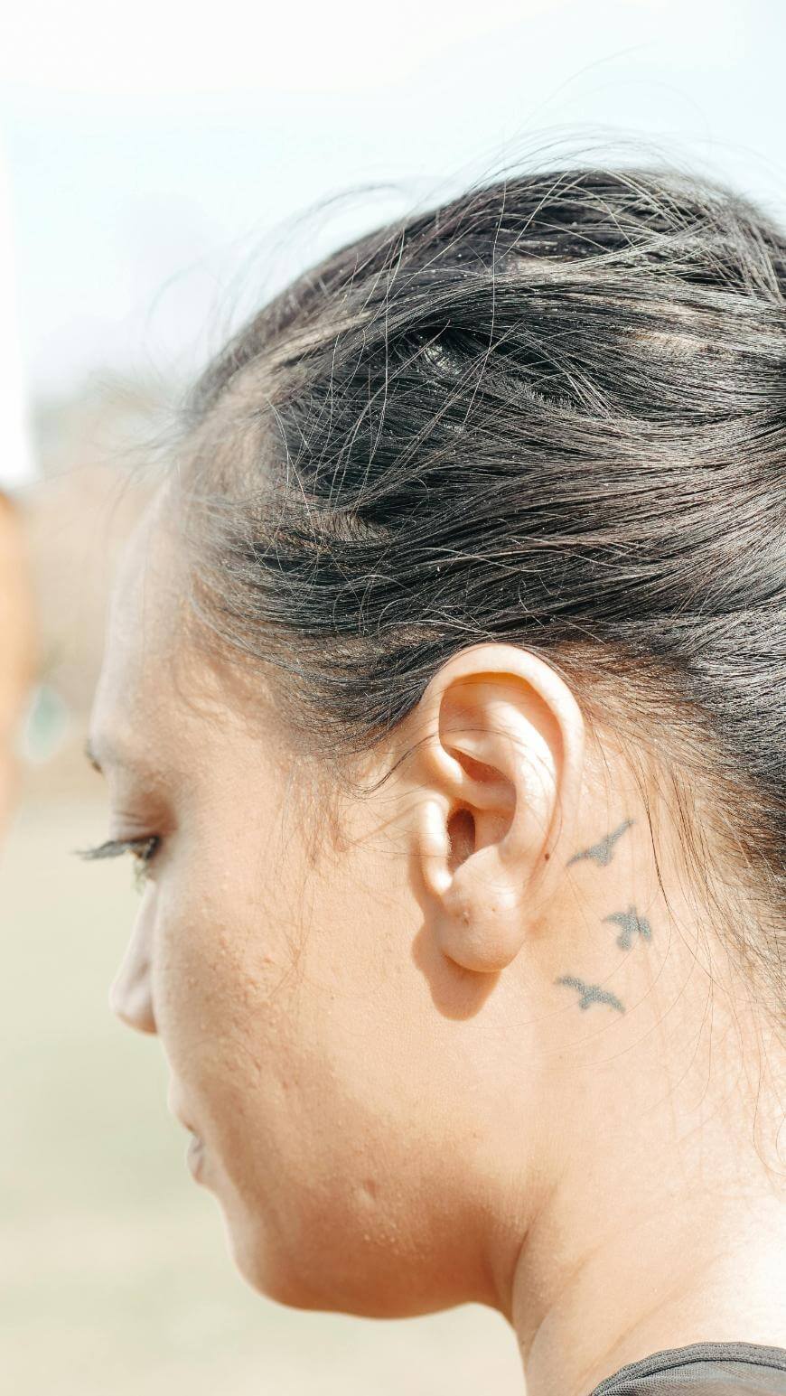 A close-up side view of a womans face and ear, showing three small bird tattoos behind her ear. She has dark hair pulled back and is looking downwards. The background is blurred.