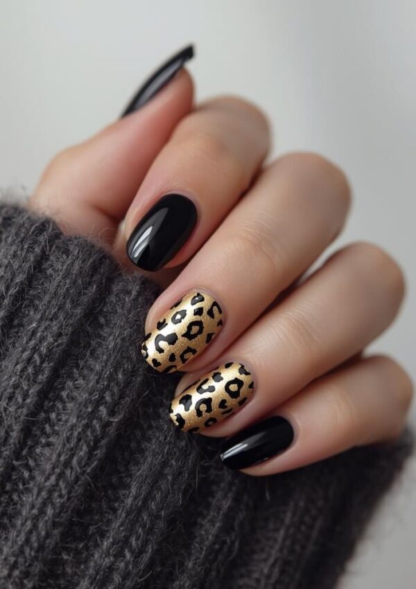 A hand with black polished nails, except for the ring and middle fingers, which have a gold base with black leopard print nail art. The hand is partially covered by a dark grey knitted sleeve.