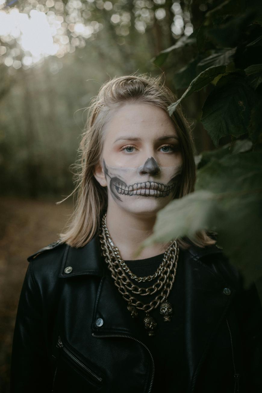 A person with long blonde hair, wearing a black leather jacket and layered chain necklaces, stands in a wooded area. Their face is painted with a black and white skull design, and leaves partially obscure the right side of their face.
