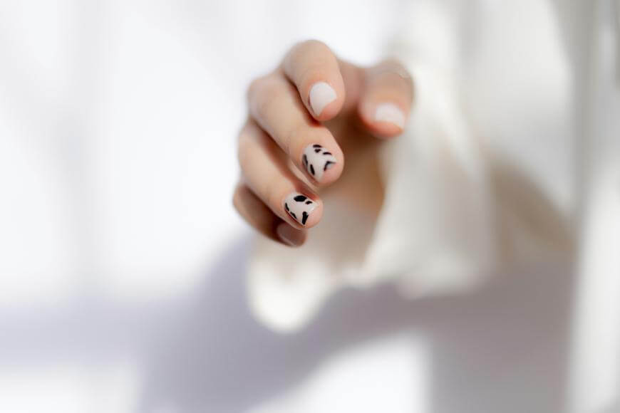 A hand with neatly manicured nails features minimalist black leaf designs on two fingernails. The person is wearing a long-sleeved white garment, and the background is softly blurred.