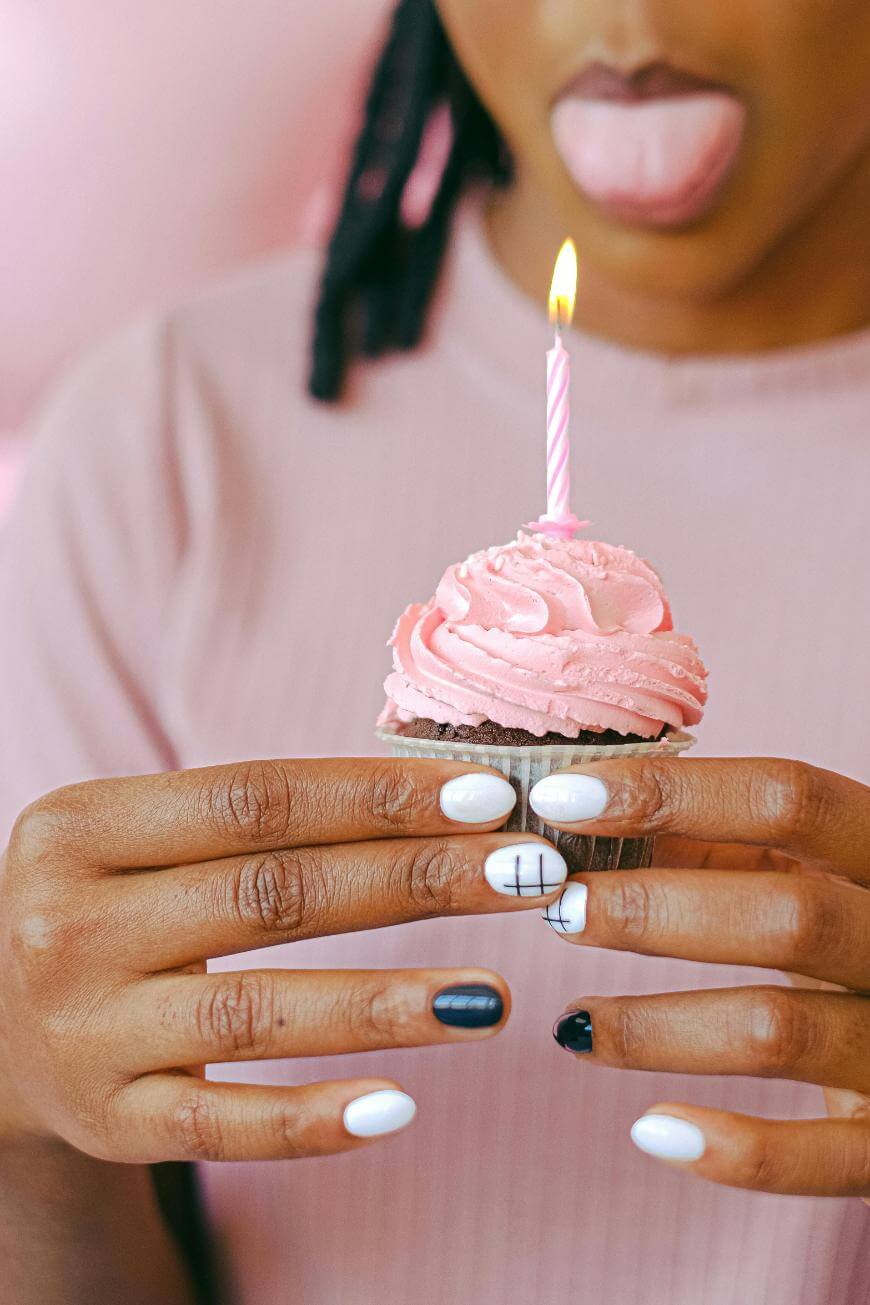 A person holds a cupcake with pink frosting and a lit candle. Their nails are painted in black, white, and pink designs. They stick out their tongue playfully and wear a light pink top.