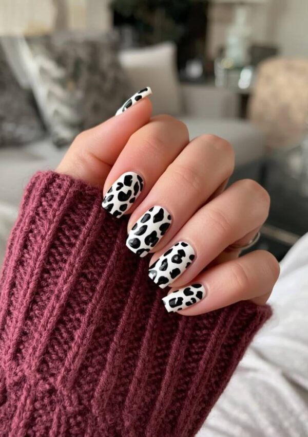 A hand with long nails painted in a black and white cow print design, partially wrapped in a maroon knit sweater sleeve, with a blurred background of a cozy room.