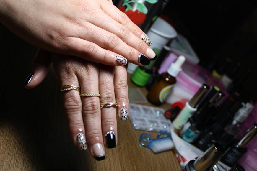 A pair of hands with leopard-print and black nail art, wearing gold rings, rests on a wooden table surrounded by nail polish bottles and manicure tools.