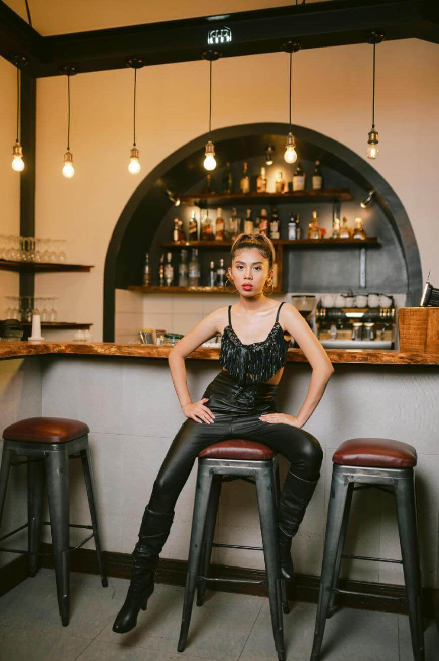 A woman in a black fringe top and leather pants sits confidently on a bar stool in a stylish bar, with shelves of bottles and hanging light bulbs in the background.