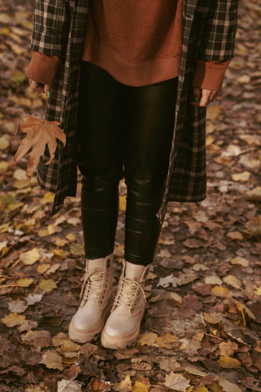 Person standing on fallen autumn leaves, wearing cream boots, black leather pants, a brown sweater, and a plaid coat, holding a large brown leaf in one hand.