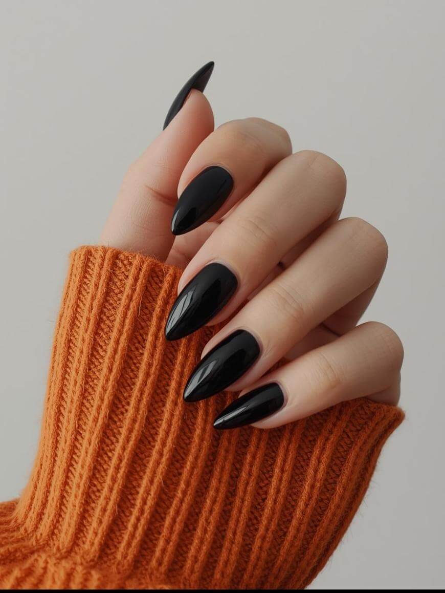 A hand with long, glossy, black stiletto-shaped nails is shown against a neutral background. The person is wearing an orange ribbed sweater with the sleeve partially covering the hand.